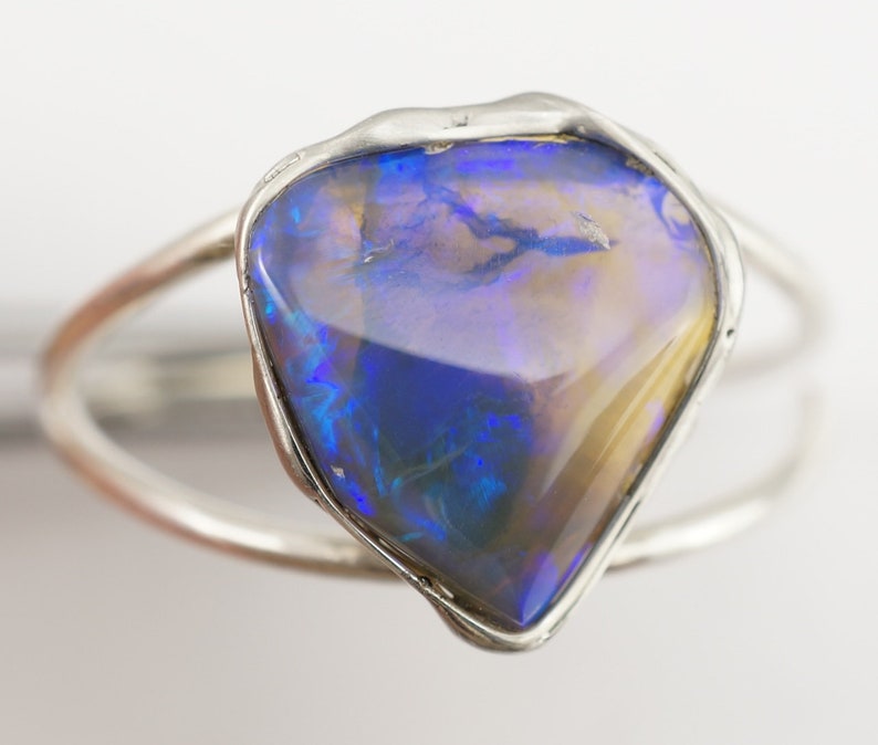 May include: A silver ring with a large, irregular shaped opal gemstone. The opal is a mix of blue, purple, and gold tones.