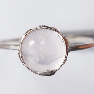 Puede incluir: Un anillo de plata con una piedra redonda, clara y rosa p&aacute;lida engastada en un delicado bisel texturizado.