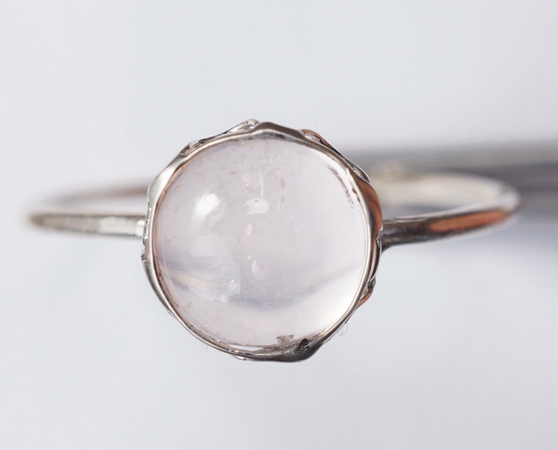 Puede incluir: Un anillo de plata con una piedra redonda, clara y rosa p&aacute;lida engastada en un delicado bisel texturizado.