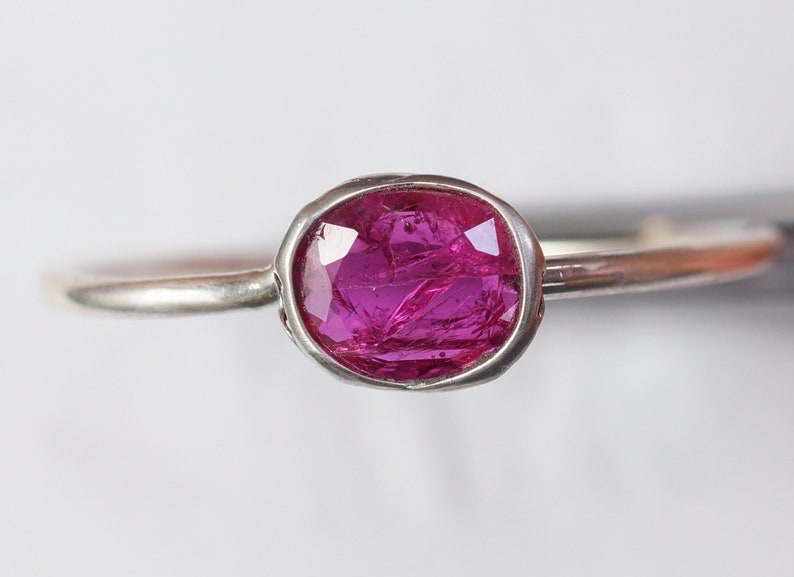 May include: A silver ring featuring a vibrant, oval-cut, magenta gemstone. The gemstone is set in a bezel setting, and the band is a simple, polished silver. The ring is displayed against a white background, highlighting the gemstone's color and clarity.