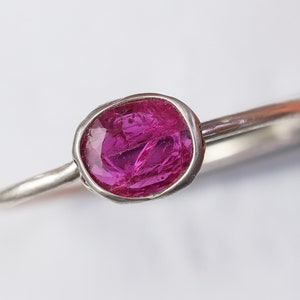May include: A close-up of a silver ring featuring a vibrant, oval-shaped, hot pink gemstone. The gemstone is set in a bezel setting, and the ring band is a simple, polished silver. The ring is set against a white background.