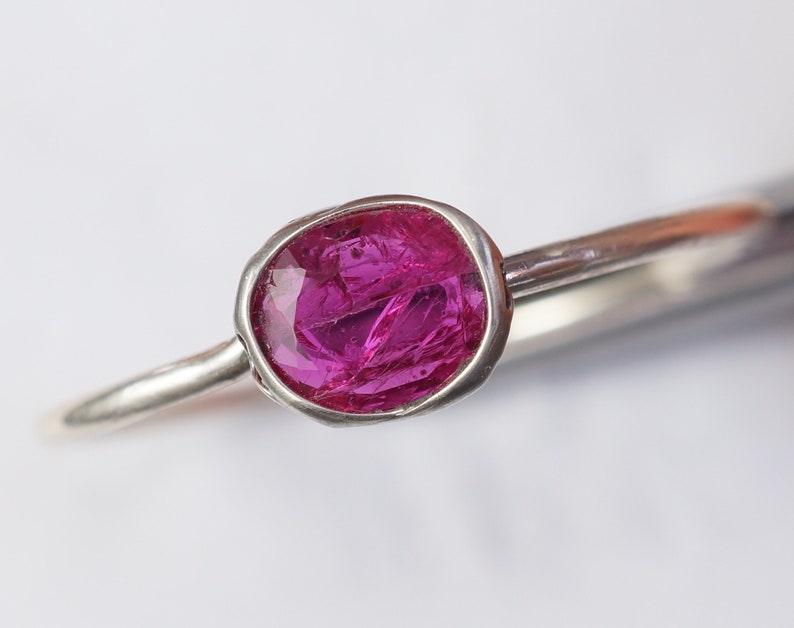 May include: A close-up of a silver ring featuring a vibrant, oval-shaped, hot pink gemstone. The gemstone is set in a bezel setting, and the ring band is a simple, polished silver. The ring is set against a white background.