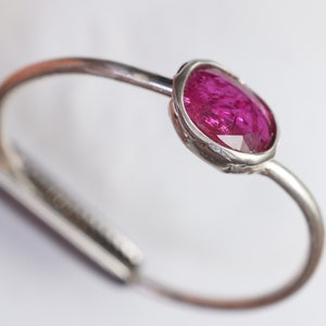 May include: A silver-toned ring featuring a vibrant, oval-cut, magenta gemstone. The gemstone is set in a bezel setting, and the ring band curves gracefully. The design is simple yet elegant, showcasing the gemstone's rich color and sparkle.