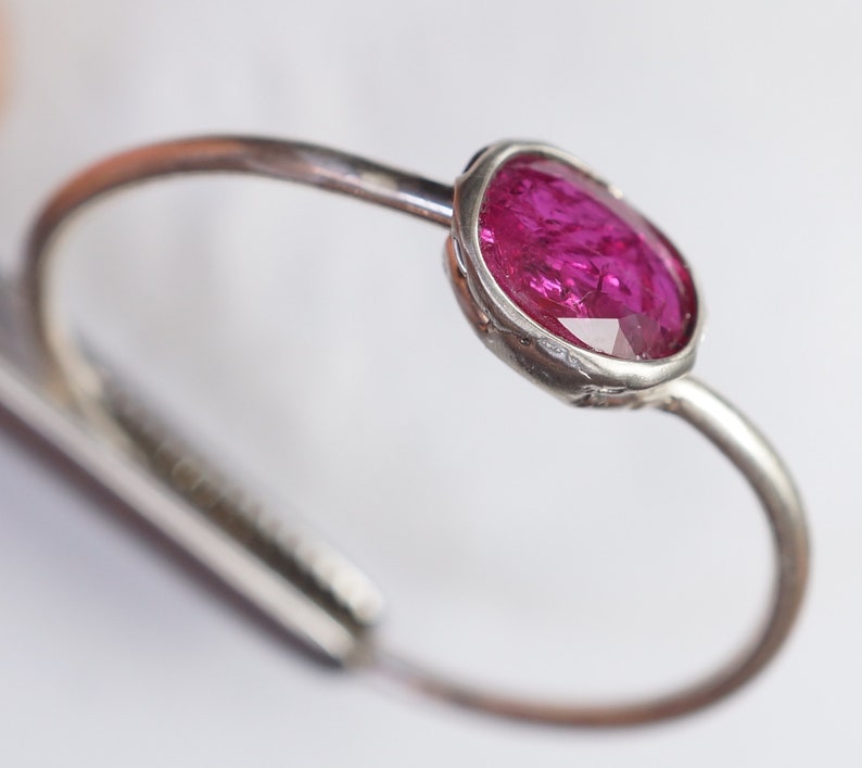 May include: A silver-toned ring featuring a vibrant, oval-cut, magenta gemstone. The gemstone is set in a bezel setting, and the ring band curves gracefully. The design is simple yet elegant, showcasing the gemstone's rich color and sparkle.