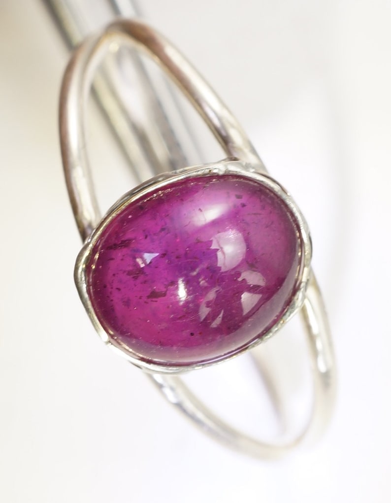 Puede incluir: Un anillo de plata con una gema grande, ovalada, de color magenta intenso. La gema est&aacute; engastada en un bisel y la banda del anillo tiene un dise&ntilde;o simple y elegante. El anillo se muestra sobre un fondo blanco.