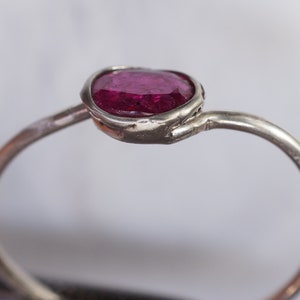 May include: A silver ring featuring a vibrant, oval-cut ruby gemstone. The ruby is set in a bezel setting, and the band has a simple, elegant design. The ring is a piece of jewelry.