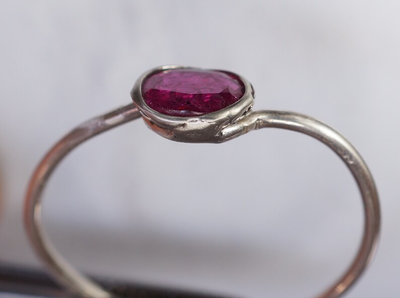 May include: A silver ring featuring a vibrant, oval-cut ruby gemstone. The ruby is set in a bezel setting, and the band has a simple, elegant design. The ring is a piece of jewelry.