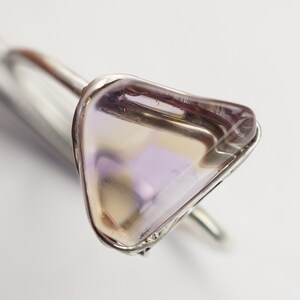 May include: A silver ring featuring a triangular amethyst gemstone. The gemstone is a pale purple colour with a touch of yellow.