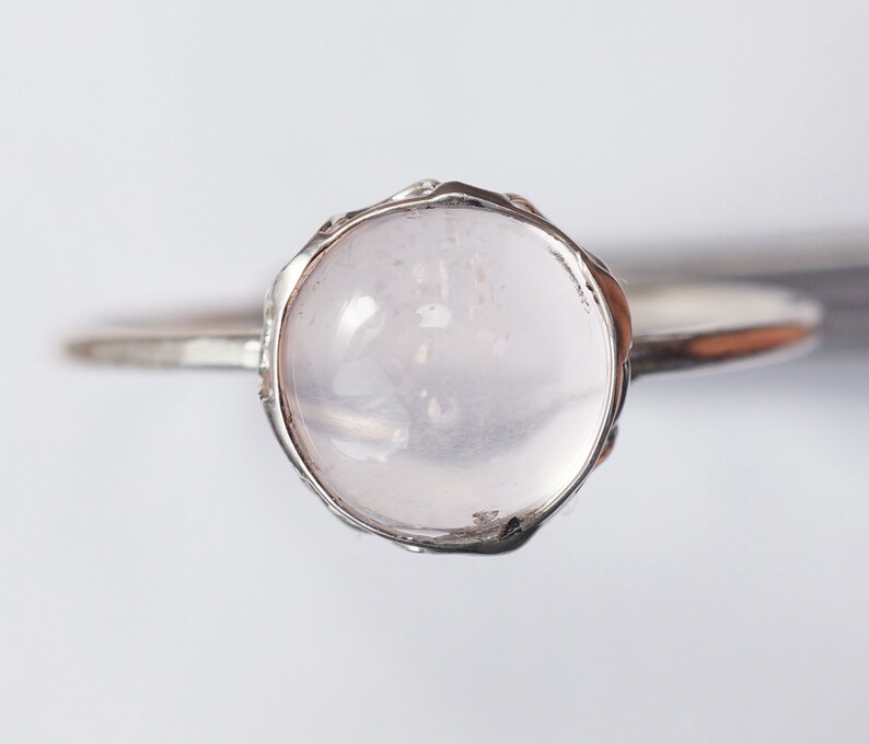 Puede incluir: Un anillo de plata con una piedra redonda, clara y rosa p&aacute;lida engastada en un engaste texturizado.
