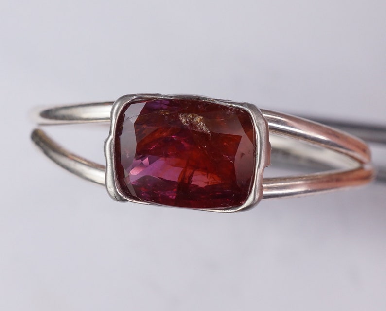 Pu&ograve; includere: Un anello d'argento con una pietra preziosa sfaccettata di colore rosso scuro incastonata in una montatura a castone.