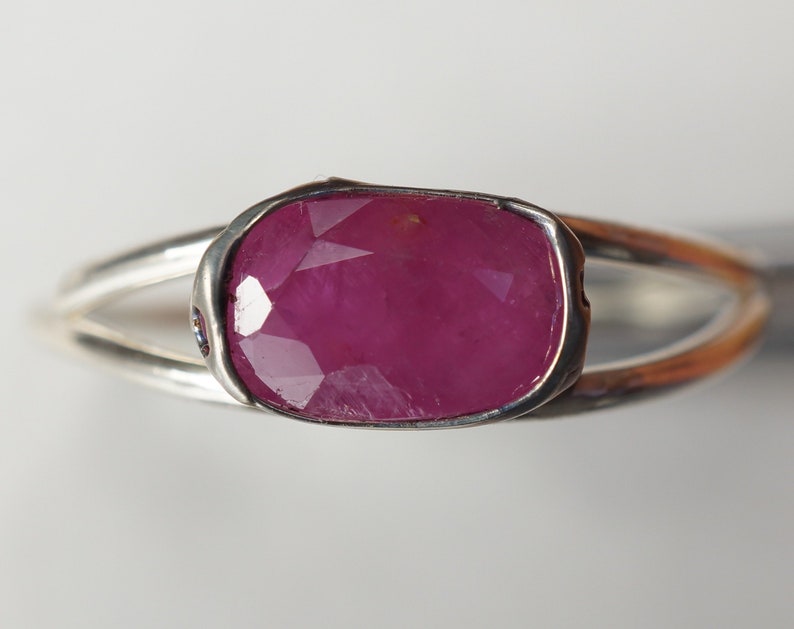 May include: A silver ring with a large, faceted, pink ruby gemstone set in a bezel setting. The ring has a split shank.