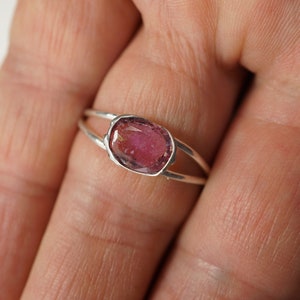 K&ouml;nnte beinhalten: Ein silberner Ring mit einem rosafarbenen Edelstein, der in einer Zargenfassung gefasst ist. Der Ring hat ein Doppelband-Design.