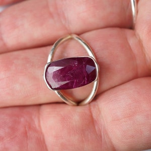 Op de afbeelding: Een zilveren ring met een grote, dieprode robijn-edelsteen gezet in een uniek ovaalvormig ontwerp.