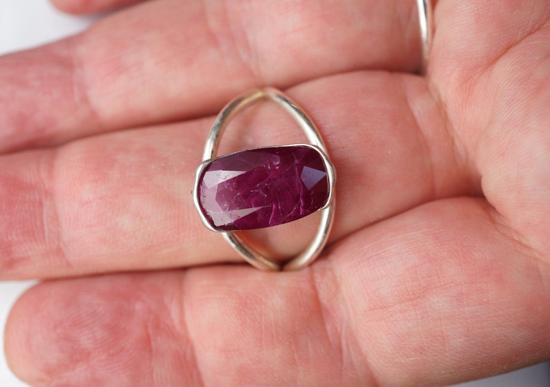 Op de afbeelding: Een zilveren ring met een grote, dieprode robijn-edelsteen gezet in een uniek ovaalvormig ontwerp.