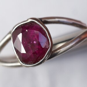 K&ouml;nnte beinhalten: Ein silberner Ring mit einem tief roten Edelstein, der in einer Zargenfassung gefasst ist. Der Ring hat ein einzigartiges, organisches Design.