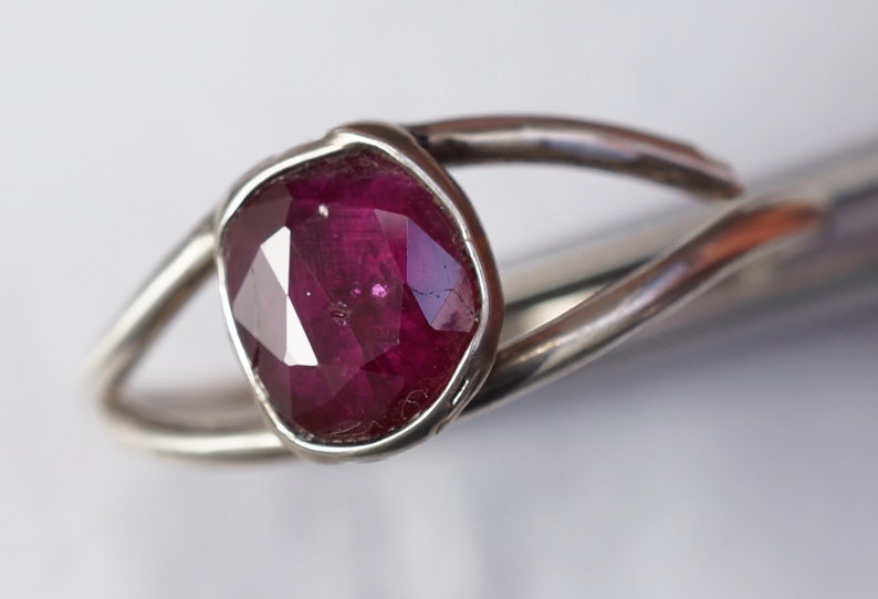 K&ouml;nnte beinhalten: Ein silberner Ring mit einem tief roten Edelstein, der in einer Zargenfassung gefasst ist. Der Ring hat ein einzigartiges, organisches Design.