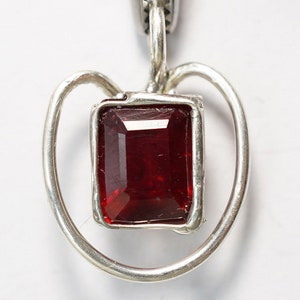Op de afbeelding: Een zilveren ketting met een hartvormige hanger en een rode edelsteen in het midden.