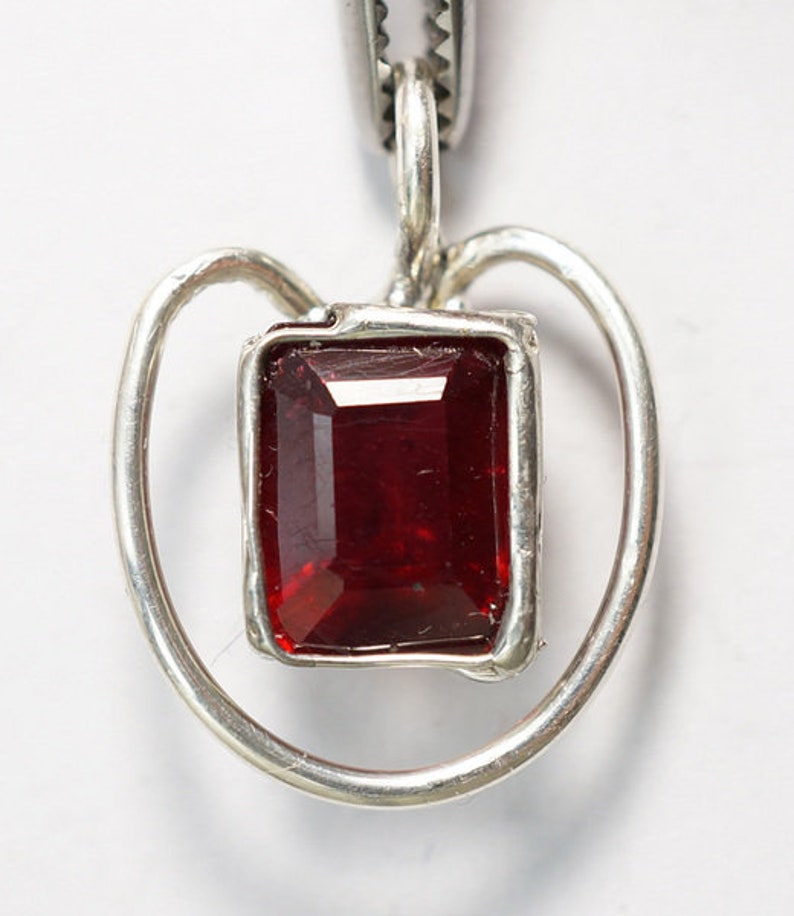 Op de afbeelding: Een zilveren ketting met een hartvormige hanger en een rode edelsteen in het midden.