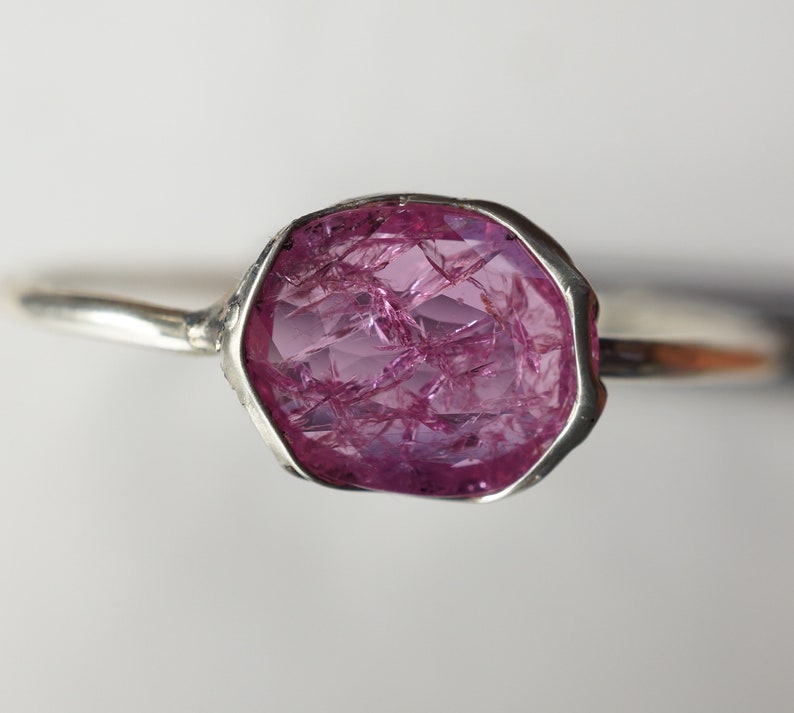 May include: A silver ring with a large, pink, faceted gemstone. The gemstone is set in a bezel setting and has a rough, natural look.