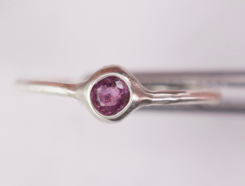 Pu&ograve; includere: Un anello d'argento con una piccola pietra preziosa rotonda di colore viola incastonata in una montatura a castone.