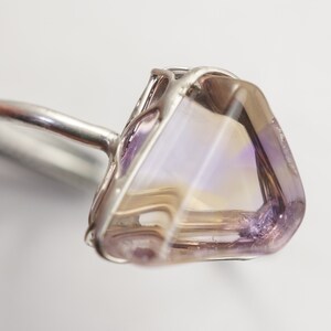 May include: A silver ring featuring a large, faceted, iridescent gemstone set in a wire cage. The gemstone displays a blend of purple, pink, and white hues.
