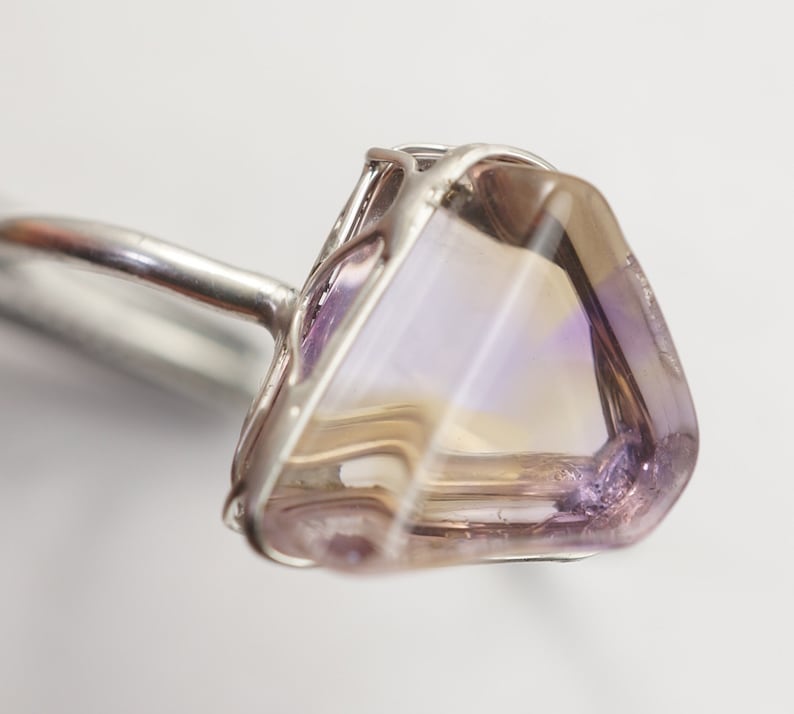 May include: A silver ring featuring a large, faceted, iridescent gemstone set in a wire cage. The gemstone displays a blend of purple, pink, and white hues.