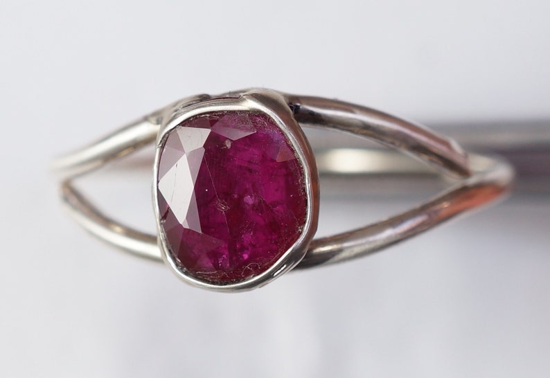 K&ouml;nnte beinhalten: Ein silberner Ring mit einem tief roten Rubin-Edelstein, der in einer Zargenfassung gefasst ist. Der Ring hat ein schlichtes, modernes Design.