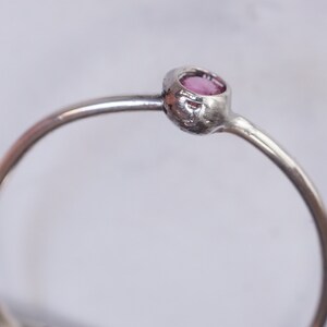 K&ouml;nnte beinhalten: Ein silberner Ring mit einem kleinen, runden, rosafarbenen Edelstein, der in einer Zargenfassung gefasst ist.