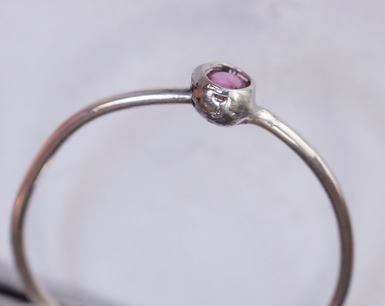 K&ouml;nnte beinhalten: Ein silberner Ring mit einem kleinen, runden, rosafarbenen Edelstein, der in einer Zargenfassung gefasst ist.