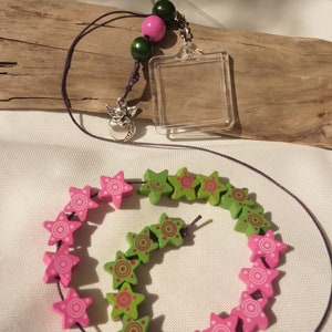 Könnte beinhalten: Eine grüne und rosafarbene, sternförmige Holzperlenkette mit einem transparenten Plastik-Bilderrahmen-Anhänger und einem silbernen Engelsanhänger. Die Kette ist auf einer dunkelvioletten Schnur aufgefädelt.