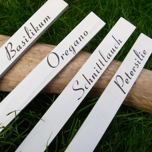 Könnte beinhalten: Vier weiße Gartenstecker aus Holz mit schwarzer Schrift. Die Stecker sind mit "Basilikum", "Oregano", "Schnittlauch" und "Petersilie" beschriftet. Die Stecker liegen auf einem Holzstamm, mit grünem Gras im Hintergrund.