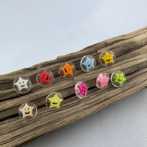 Könnte beinhalten: Sortiment von zehn sternförmigen Knöpfen mit Smiley-Gesichtern. Die Knöpfe sind aus klarem Kunststoff mit verschiedenen farbigen Smiley-Gesichtern im Inneren. Die Knöpfe sind auf einem Stück Treibholz angeordnet.
