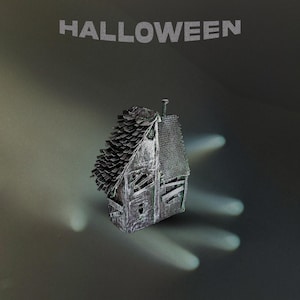 Puede incluir: Un modelo en miniatura de una casa en ruinas con un acabado oscuro y desgastado, que presenta un techo texturizado y ventanas tapiadas. La palabra HALLOWEEN está impresa en letras mayúsculas grandes y claras sobre la estructura en un fondo oscuro.