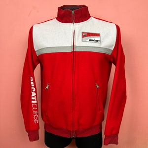 May include: Red fleece jacket with white and gray accents, featuring a full-length zipper and a high collar. The jacket has the word "DUCATI" on a patch and "DUCATI CORSE" printed on the sleeve. Two zippered pockets are visible.