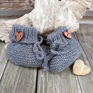 May include: A pair of gray knitted baby booties with wooden heart-shaped buttons. The booties are on a wooden surface.