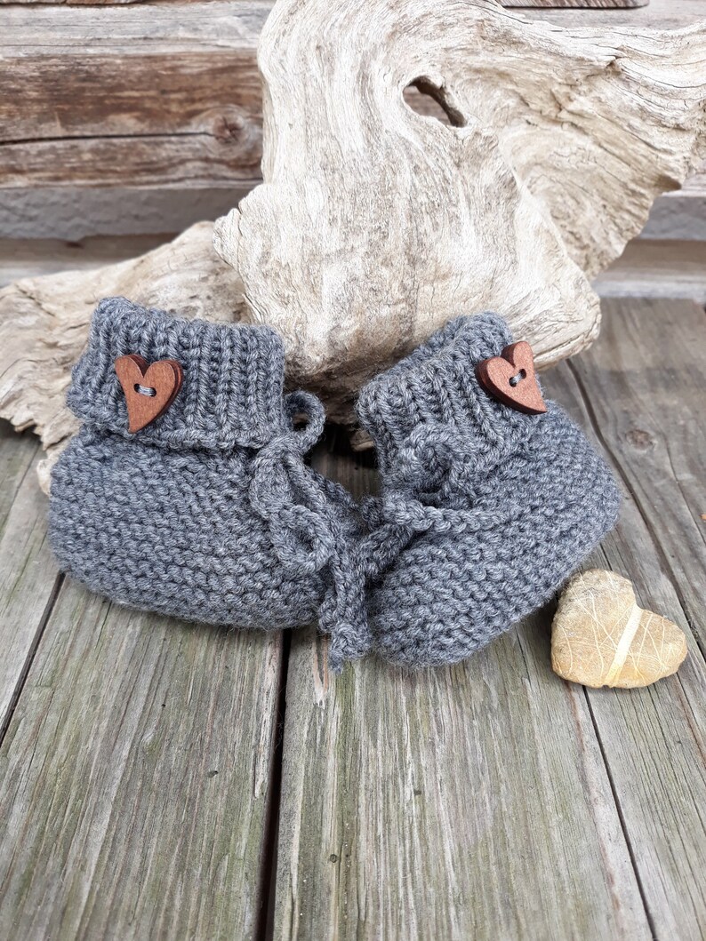 May include: A pair of gray knitted baby booties with wooden heart-shaped buttons. The booties are on a wooden surface.