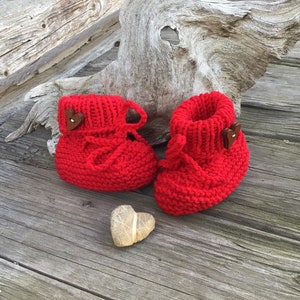 May include: A pair of red knitted baby booties with brown heart-shaped buttons. The booties are on a wooden surface with a heart-shaped rock.