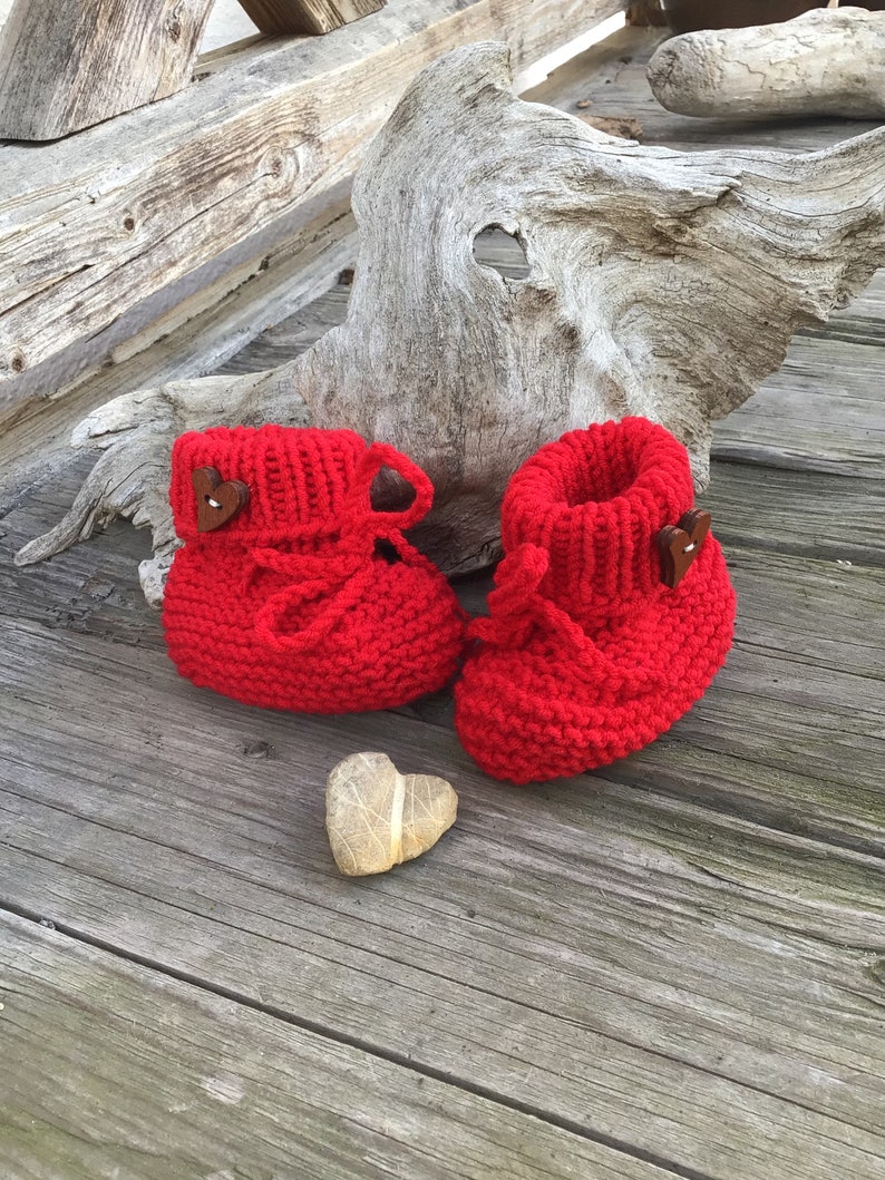 May include: A pair of red knitted baby booties with brown heart-shaped buttons. The booties are on a wooden surface with a heart-shaped rock.