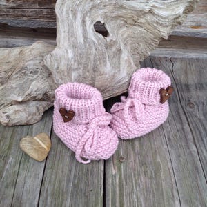 May include: A pair of pink knitted baby booties with brown heart-shaped buttons. The booties are tied with pink ribbon laces.