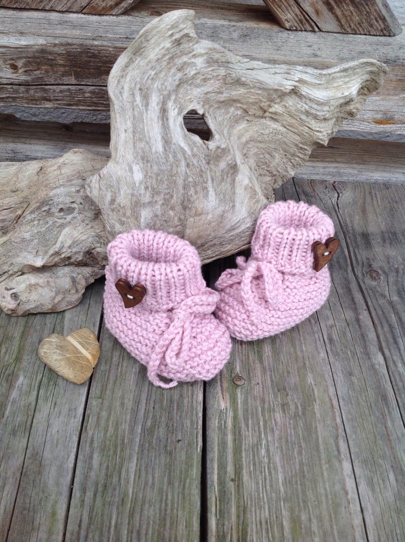 May include: A pair of pink knitted baby booties with brown heart-shaped buttons. The booties are tied with pink ribbon laces.