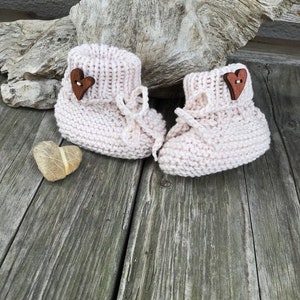 May include: A pair of pink knitted baby booties with wooden heart buttons. The booties are on a wooden surface.