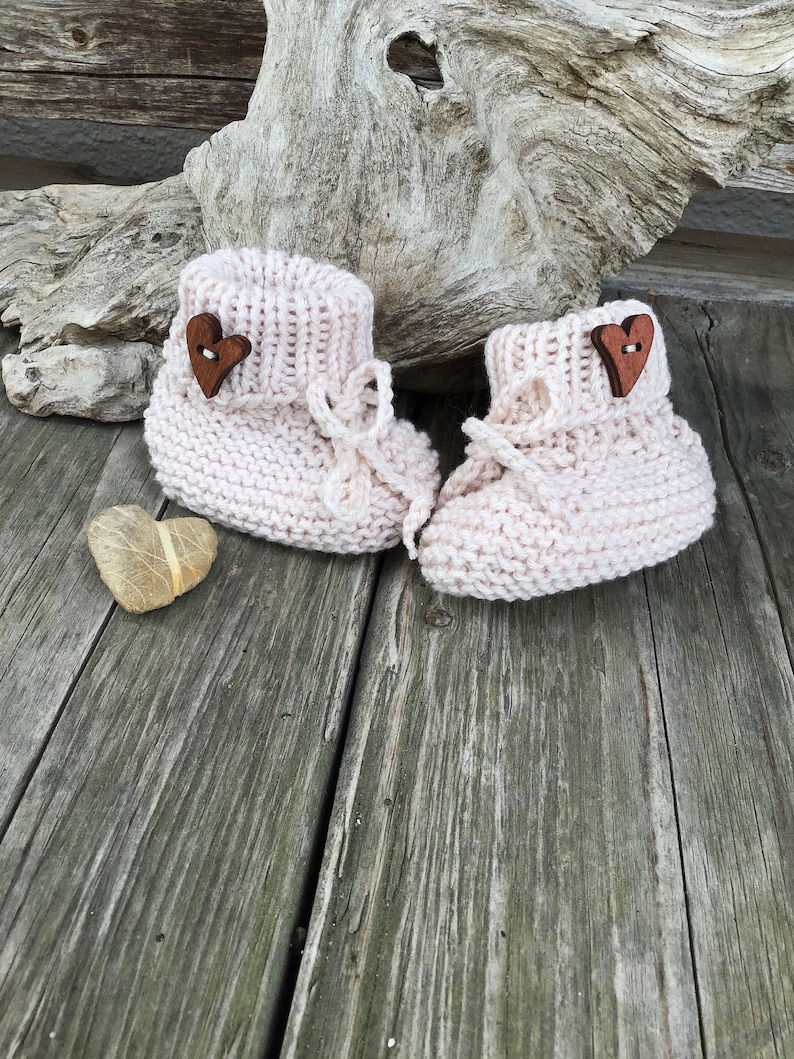 May include: A pair of pink knitted baby booties with wooden heart buttons. The booties are on a wooden surface.