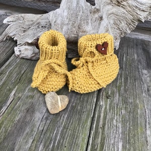 May include: A pair of yellow knitted baby booties with a brown heart button on one bootie. The booties are sitting on a wooden surface.