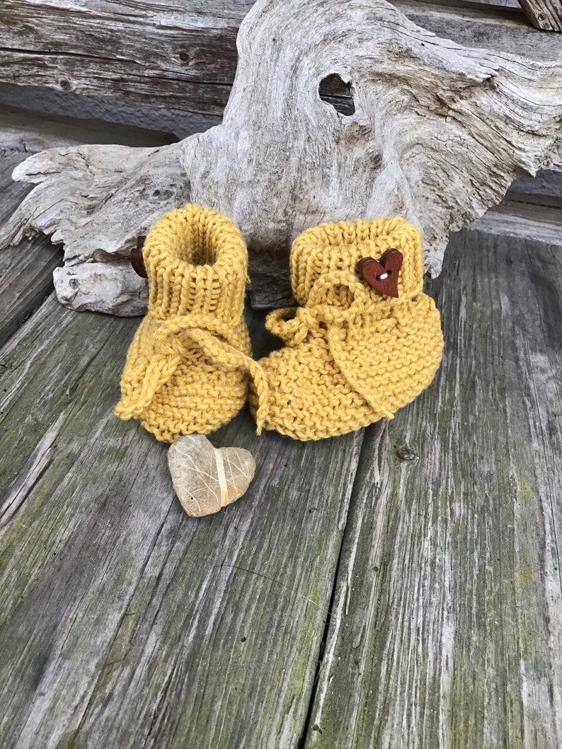 May include: A pair of yellow knitted baby booties with a brown heart button on one bootie. The booties are sitting on a wooden surface.