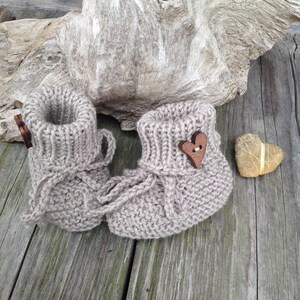May include: A pair of beige knitted baby booties with a wooden heart button on one bootie. The booties are sitting on a wooden surface.