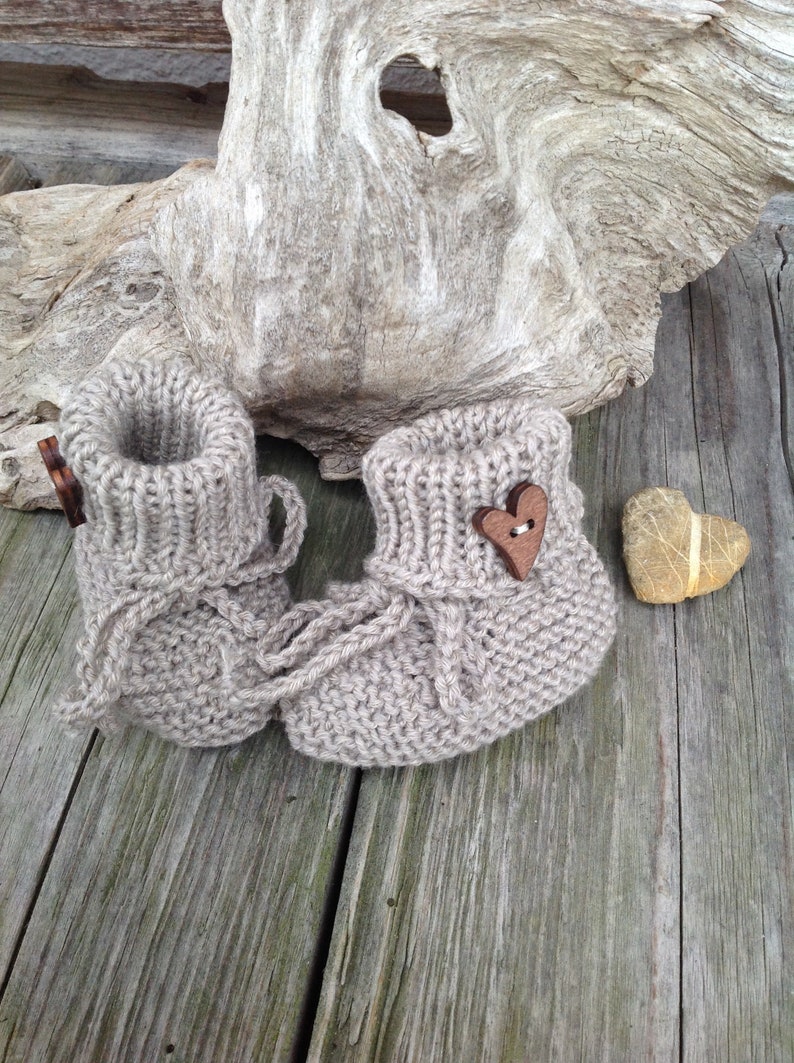 May include: A pair of beige knitted baby booties with a wooden heart button on one bootie. The booties are sitting on a wooden surface.