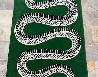 May include: A rectangular green rug featuring a large, stylized snake design. The snake is depicted in black and white, with a detailed pattern. The snake's head has a red tongue and a small leaf. The rug is placed on a stone floor.