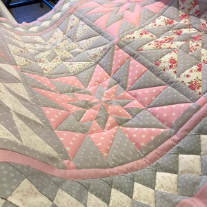 Könnte beinhalten: Eine handgefertigte Steppdecke mit einem Sternmuster in Rosa, Grau und Weiß. Das Design umfasst Punkte und florale Muster innerhalb geometrischer Formen. Die Kanten sind mit einem rosa Rand versehen.