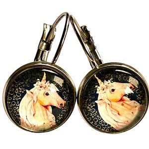 Könnte beinhalten: Ein Paar silberne Creolen mit einem runden Glasanhänger, der ein pinkfarbenes Einhorn mit einer Blumenkrone auf einem dunkelblauen Hintergrund mit goldenem Glitzer zeigt.