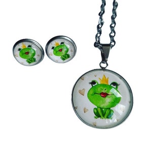 Könnte beinhalten: Ein silbernes Halsketten- und Ohrring-Set mit einem grünen Frosch, der eine goldene Krone und eine rote Lippe trägt. Der Frosch ist von weißen Herzen auf weißem Hintergrund umgeben.